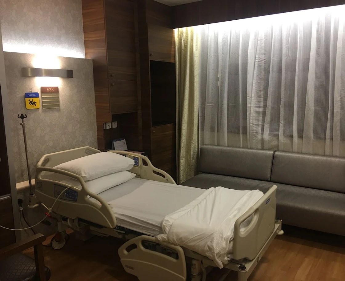 An adjustable hospital bed with two pillows and a slightly raised head section beside a gray sofa